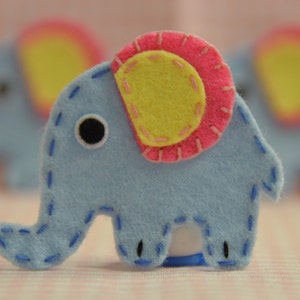 May include: A light blue felt elephant with a pink and yellow ear design. The elephant has a black eye and is stitched with blue thread. The image shows three elephants, with the main one in focus.