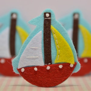 May include: A felt sailboat with a red hull, white sails, and a yellow sail. The boat has a light blue background and a brown mast. White dots decorate the hull. The image shows multiple sailboats.
