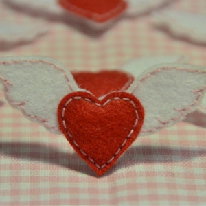 Può includere: Primo piano di diversi cuori di feltro rosso con ali di feltro bianco, cuciti con filo rosa. I cuori sono su un tessuto a quadretti rosa e bianco. Le ali sono attaccate ai cuori.