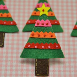 May include: Handmade felt Christmas tree ornaments. Each tree is green with colorful orange, pink, and red accents. The trees have brown trunks and are topped with a yellow star. The background is a pink and white checkered pattern.