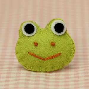 Könnte beinhalten: Ein handgefertigtes, hellgrünes Froschgesicht aus Filz mit schwarzen Augen, weißen Augenringen und einem orangefarbenen, aufgestickten Lächeln. Der Frosch ist ein skurriles Design, perfekt für Bastelarbeiten oder als dekoratives Element. Der Hintergrund ist ein rosa-weißes Karomuster.