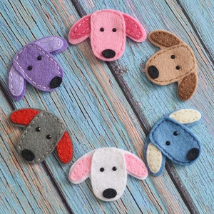 Op de afbeelding: Zes vilten hondenkoppen in verschillende kleuren, waaronder roze, paars, bruin, grijs, wit en blauw. Elke hond heeft een zwarte neus en ogen, met contrasterende oorkleuren. De vilten honden zijn in een cirkelvormig patroon op een blauw houten oppervlak gerangschikt.