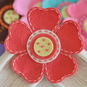 Puede incluir: Primer plano de una flor de fieltro hecha a mano con pétalos rojos y un contorno cosido blanco. El centro presenta un círculo amarillo con pequeños puntos rosas. Otras flores de fieltro de varios colores están en el fondo.