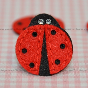 May include: A red felt ladybug with black spots and a black head. The ladybug has two clear rhinestones for eyes. The image is of a craft item, possibly a button or embellishment. The text "Designed by ASEC Inc." is visible.