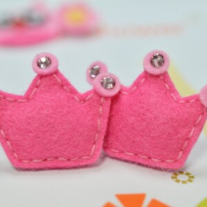 Puede incluir: Dos coronas de fieltro rosa brillante con pedrería plateada. Las coronas están cosidas con hilo rosa claro y tienen un diseño sencillo. Perfectas para manualidades o adornos, estas coronas añaden un toque de fantasía.