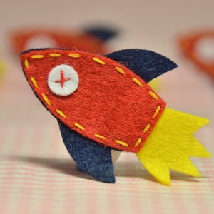 May include: A handmade felt rocket ship in red, navy blue, and yellow. The rocket has a white button with a red cross and yellow stitching. The rocket is approximately 3 inches long.
