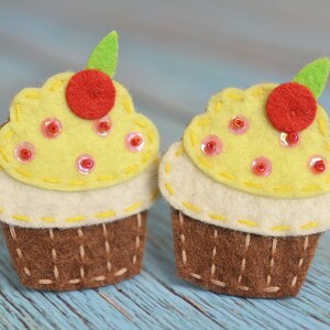 Op de afbeelding: Twee handgemaakte vilten cupcakes. Elke cupcake heeft een bruine basis, een crèmekleurige laag en een gele bovenkant versierd met rode pailletten en een rode kers met een groen blad. De cupcakes staan op een lichtblauwe houten ondergrond.