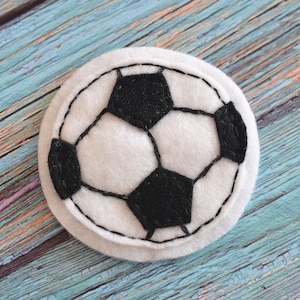 May include: A round, white felt coaster with a black and white soccer ball design. The soccer ball pattern is created with black felt and stitched onto the white felt base. The coaster is placed on a weathered, blue wooden surface.