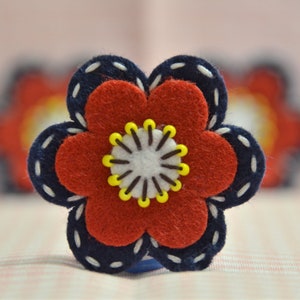 May include: A handmade felt flower with a red centre, navy blue petals, and white stitching. The flower has a white and yellow centre. The background is a soft pink.