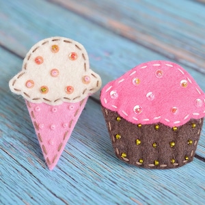 Puede incluir: Dos elementos de comida de fieltro: un cono de helado y una magdalena. El cono de helado tiene un cono rosa y una bola de helado crema con cuentas rosas y doradas. La magdalena tiene una base marrón y una parte superior rosa con cuentas doradas. Ambos artículos tienen detalles cosidos.