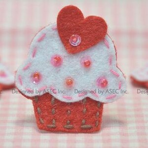 Puede incluir: Decoración en forma de cupcake de fieltro, con base roja y parte superior blanca, adornada con un corazón rojo y cuentas rosas. El diseño incluye detalles cosidos y el texto "Designed by ASEC Inc."