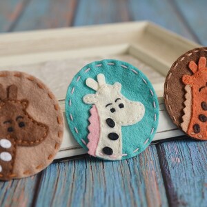 May include: Three felt giraffe patches in brown, teal, and tan. Each patch features a giraffe head with pink, white, and brown accents. The patches are round with stitched edges, perfect for crafting or embellishing.