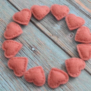 May include: A collection of small, heart-shaped felt ornaments in a muted rose color, arranged in a circle. Each heart is hand-stitched with a contrasting thread, set against a weathered, blue-toned wooden surface. Ideal for crafts and decorations.