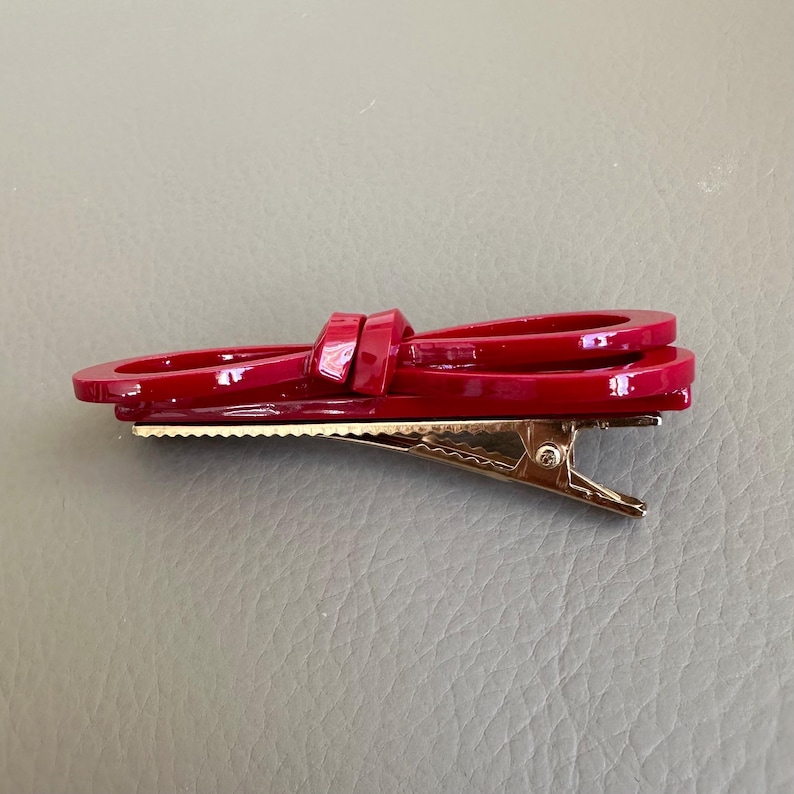 May include: A glossy red hair clip in a bow shape. The clip has a gold-colored metal clasp. The bow is made of a shiny, solid red material. The hair accessory is designed to hold hair in place.