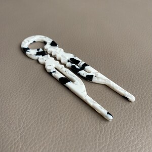 May include: Two white hair pins with a black speckled pattern. The pins have a unique design with a loop at the top and a forked end. The pins are made of a glossy material and are laying on a neutral-toned surface.