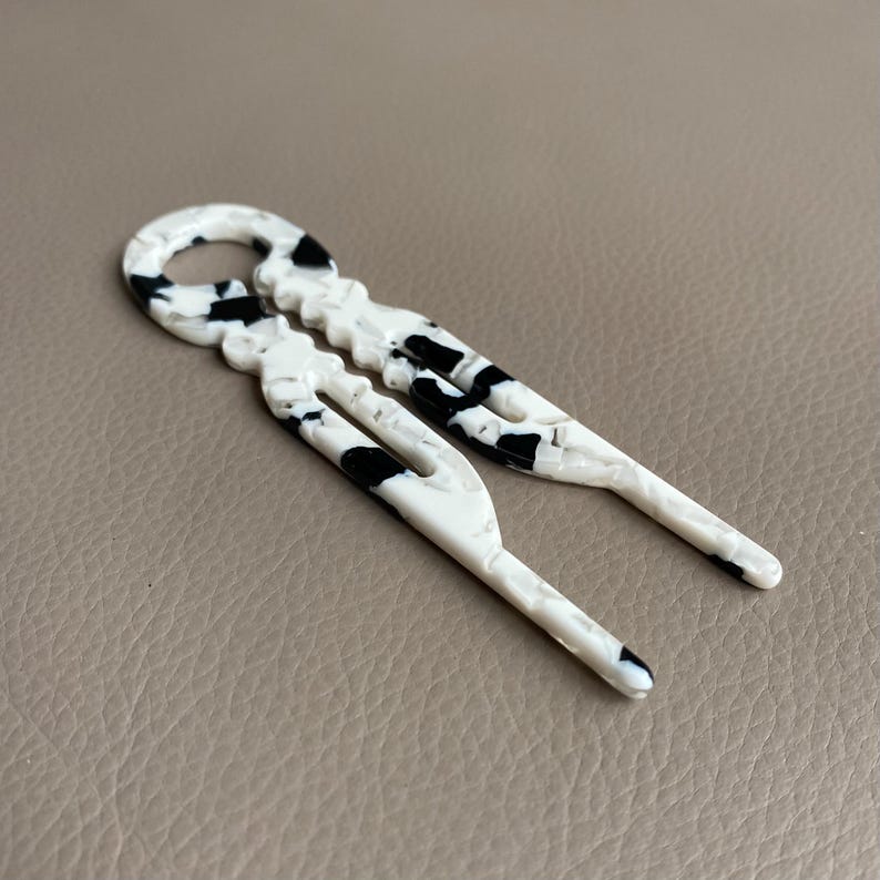May include: Two white hair pins with a black speckled pattern. The pins have a unique design with a loop at the top and a forked end. The pins are made of a glossy material and are laying on a neutral-toned surface.