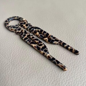 May include: Two hair pins with a tortoiseshell pattern. The pins are brown, black, and tan, with a glossy finish. The pins have a loop at the top and a forked design.