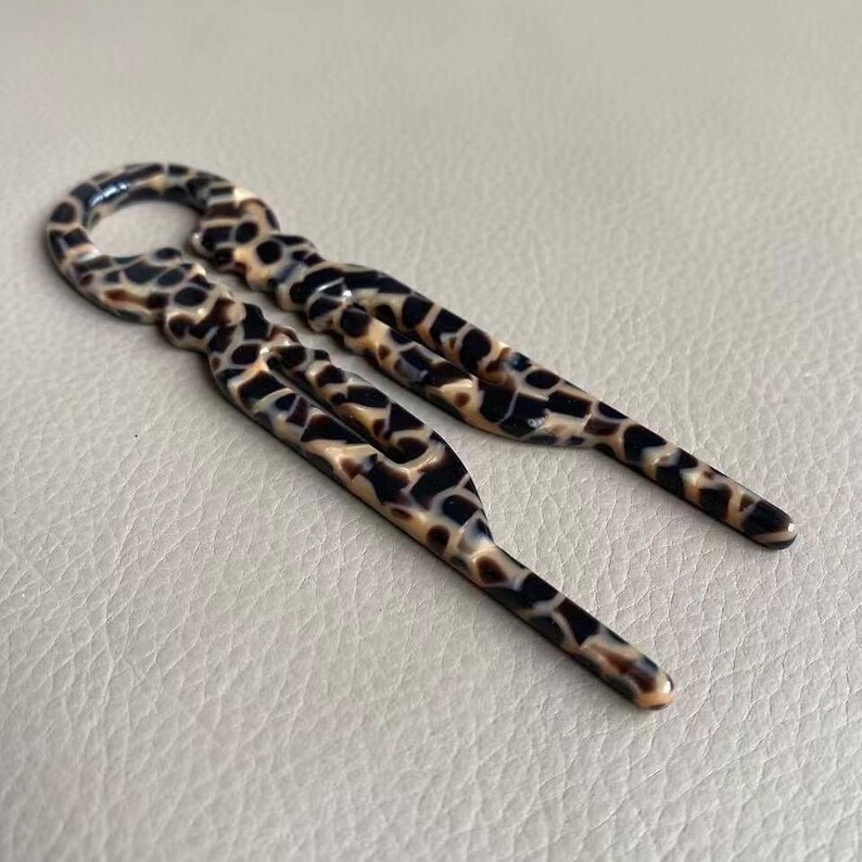 May include: Two hair pins with a tortoiseshell pattern. The pins are brown, black, and tan, with a glossy finish. The pins have a loop at the top and a forked design.