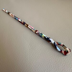 May include: A colorful hair stick with a swirling design and an oval-shaped opening. The stick features a mix of colors, including pink, white, brown, and red. The hair accessory is made of a glossy material.