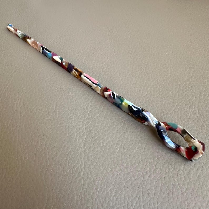 May include: A colorful hair stick with a swirling design and an oval-shaped opening. The stick features a mix of colors, including pink, white, brown, and red. The hair accessory is made of a glossy material.