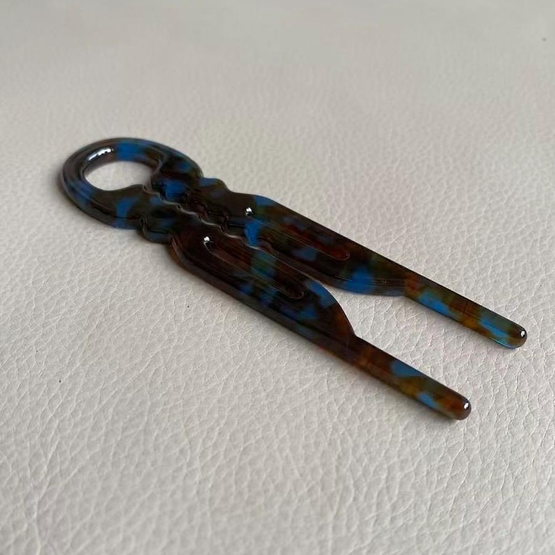 May include: A decorative hair accessory with a unique design. The hair clip features a marbled pattern of brown, blue, and amber hues. The accessory has a loop at the top and two prongs extending downward.