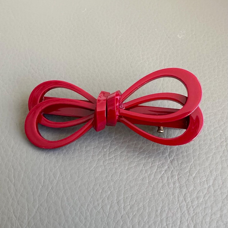 May include: A bright red bow-shaped hair clip. The clip is made of a glossy material and features a classic bow design. The hair accessory is a vibrant shade of red.