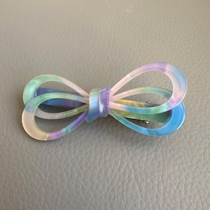 May include: A pastel-colored bow hair clip. The clip features a bow shape with a gradient of colors, including blue, green, purple, and yellow. The bow is made of a translucent material and has a metal clip on the back.