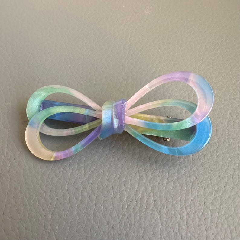 May include: A pastel-colored bow hair clip. The clip features a bow shape with a gradient of colors, including blue, green, purple, and yellow. The bow is made of a translucent material and has a metal clip on the back.