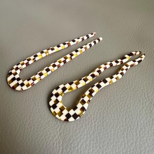 Könnte beinhalten: Zwei karierte Haargabeln. Die Haaraccessoires weisen ein Muster aus weißen, braunen und gelben Quadraten auf. Die Haargabeln sind gebogen und haben oben zwei Zinken. Der Hintergrund ist einfarbig grau.