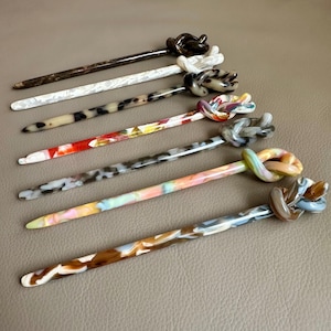 Puede incluir: Colección de siete horquillas para el cabello con la parte superior en forma de nudo. Las horquillas presentan varios colores y patrones, incluyendo marrón, blanco, carey, rojo y diseños multicolores. Los accesorios para el cabello están dispuestos sobre una superficie de tono neutro.