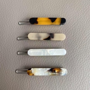 May include: Four rectangular hair clips with metal clasps. The clips have a variety of patterns, including a tortoiseshell design, a marbled pattern, and a pearlescent white. The clips are designed for securing hair.