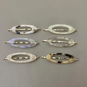 Puede incluir: Seis horquillas ovaladas para el cabello con detalles metálicos dorados. Las horquillas tienen un diseño veteado en varios colores, incluyendo blanco, gris y marrón. Los accesorios para el cabello están dispuestos sobre una superficie gris.