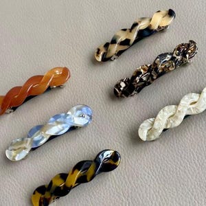 May include: A collection of six wavy hair barrettes in various colors and patterns. The barrettes feature a twisted design and are made of a glossy material. Colors include tortoiseshell, amber, cream, and blue.