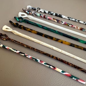 May include: An assortment of colorful hair sticks in various patterns and colors. The sticks feature a variety of designs, including swirls, knots, and loops. Colors include white, black, green, brown, and multi-colored patterns.