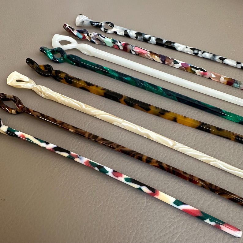 May include: An assortment of colorful hair sticks in various patterns and colors. The sticks feature a variety of designs, including swirls, knots, and loops. Colors include white, black, green, brown, and multi-colored patterns.