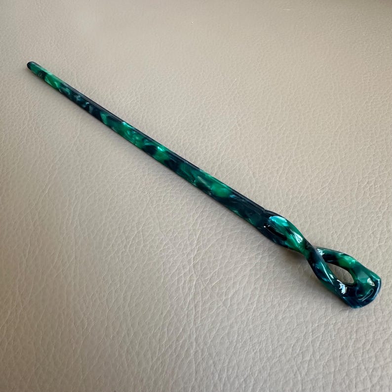 May include: A green and blue marbled hair stick with a twisted design. The hair accessory is long and slender, with a pointed end and a looped end. The color pattern is a mix of dark green and blue, creating a unique look.