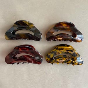 May include: Four claw hair clips in various colors. The clips have a tortoiseshell pattern in shades of brown, amber, and black. Each clip has a curved design with a central opening and small teeth for grip.