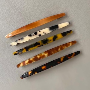 May include: A set of five long, slender hair barrettes arranged diagonally. The top clip is solid amber, followed by a white clip with black spots, a yellow and black tortoiseshell clip, a brown tortoiseshell clip, and a dark brown tortoiseshell clip.