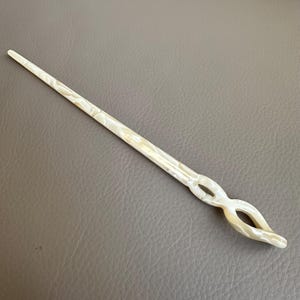 May include: A pearlescent white hair stick with a decorative openwork design. The hair accessory is long and slender, with a pointed end and a curved, looped design near the top. The stick is approximately 7 inches long.