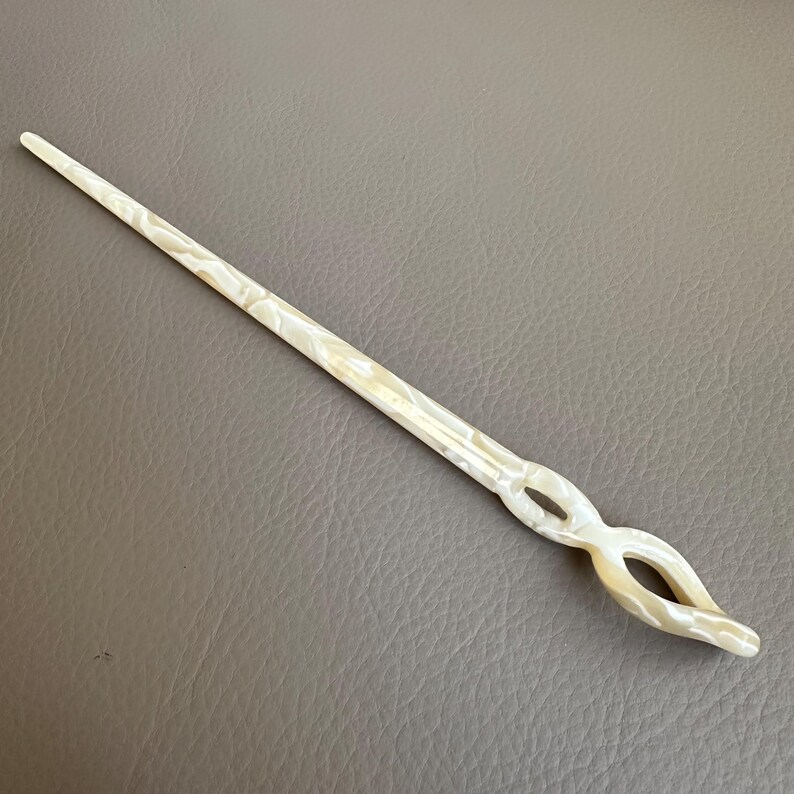 May include: A pearlescent white hair stick with a decorative openwork design. The hair accessory is long and slender, with a pointed end and a curved, looped design near the top. The stick is approximately 7 inches long.