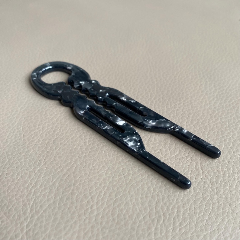 May include: Two dark blue hair barrettes with a marbled white pattern. Each barrette has a loop at the top and a three-pronged design. The barrettes are made of a glossy material.