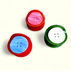 May include: Three colorful buttons with four holes each. One button is red with a light blue center, another is red with a pink center, and the third is green with a white center. The buttons are on a white background.