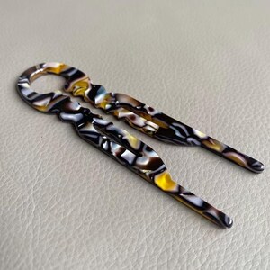 May include: Two decorative hair pins with a marbled pattern in shades of yellow, brown, black, and white. The pins have a loop at the top and a pointed end. The pins are designed for securing hair in place.