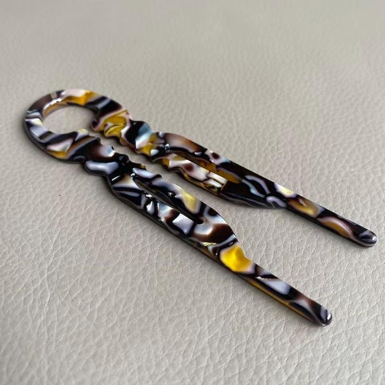 May include: Two decorative hair pins with a marbled pattern in shades of yellow, brown, black, and white. The pins have a loop at the top and a pointed end. The pins are designed for securing hair in place.