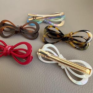 May include: Five bow-shaped hair clips in various colors. The clips include a red, brown, tortoiseshell, iridescent, and white clip. Each clip has a gold-colored metal clasp.
