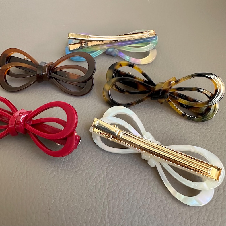 May include: Five bow-shaped hair clips in various colors. The clips include a red, brown, tortoiseshell, iridescent, and white clip. Each clip has a gold-colored metal clasp.