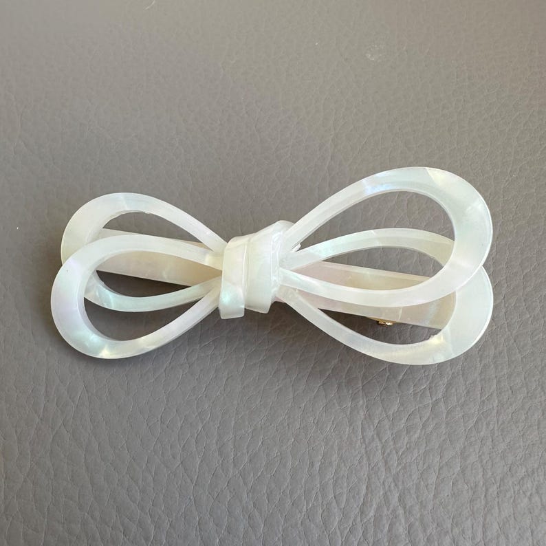 May include: A pearlescent white bow hair clip. The clip is in the shape of a bow with two loops on each side and a knot in the center. The clip is made of a shiny, iridescent material and has a gold-colored clasp.