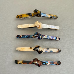 May include: Five hair clips with knotted designs. The clips are made of a tortoiseshell-like material with different color variations. The clips are arranged vertically in a row.