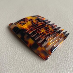 Puede incluir: Un peine para el cabello con estampado de carey, con una parte superior curvada y m&uacute;ltiples dientes. El peine presenta una mezcla de tonos marrones, negros y &aacute;mbar, creando un accesorio cl&aacute;sico y elegante. Los dientes est&aacute;n espaciados y puntiagudos.