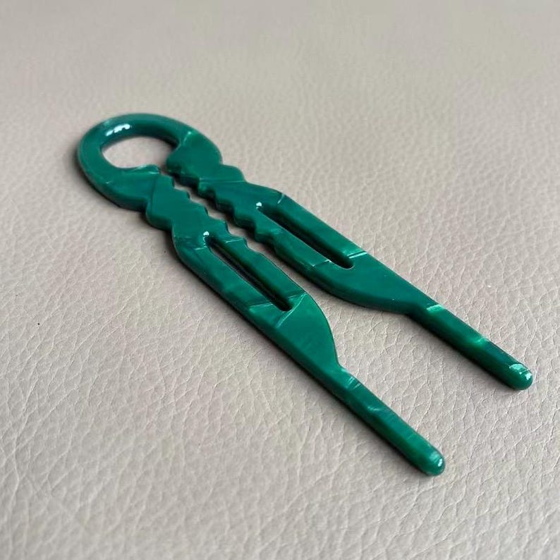 May include: A pair of green hair clips with a marbled effect. The clips are made of a glossy material and have a unique, open design with a loop at the top. The two sides of the clip are connected at the top.